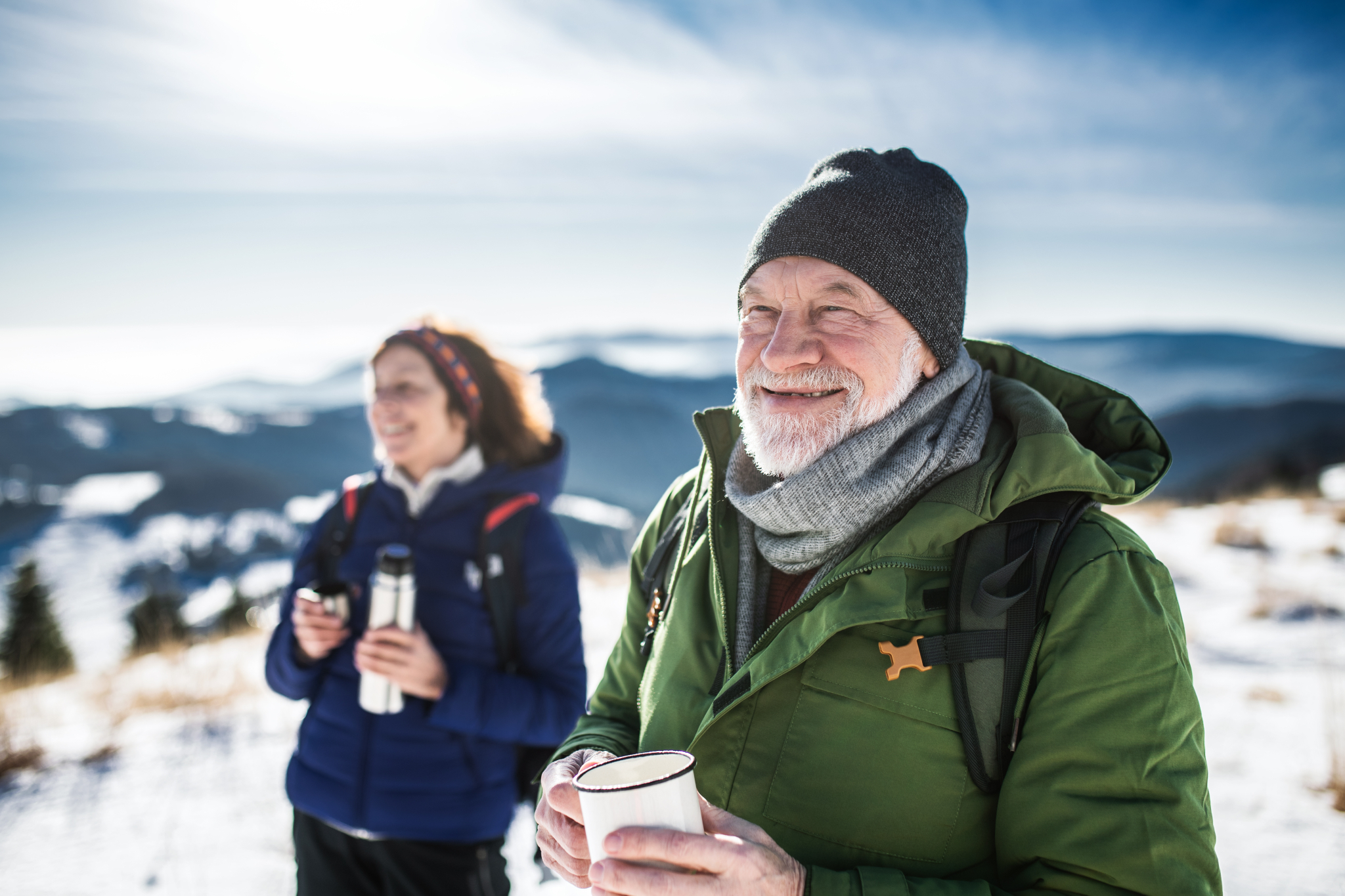 Eldre ektepar på en fjelltopp en vinterdag med en varm kopp drikke i hendene.